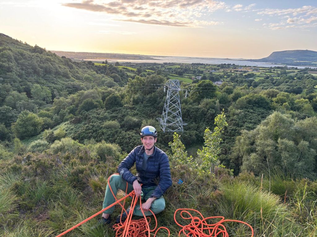 Otis Sitting on a hilltop dressed in climbing gear with a rope in the foreground.  behind him is a wide view of bush  and then a large bay.