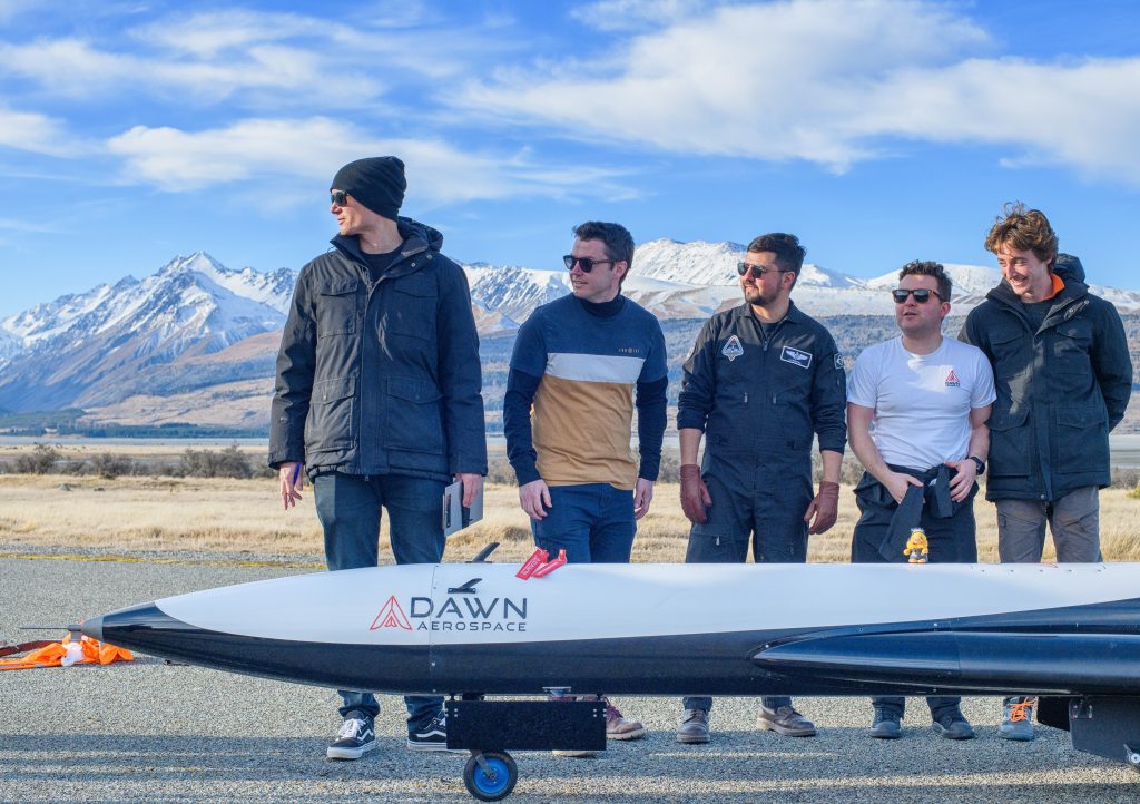 Otis and workmates standing in a line behind a Dawn Aerospace spaceplane on a runway.  Otis is on the far left of the group wearing a hat.