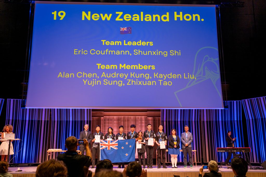 Picture =of the NZ rep team on stage after presentation of their honourable mention medals. The team are wearing team shirts and their medals. They and teh team leaders are standing in a line under a slide announcing their 19th place.
